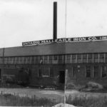 Black and white photo of a large two storey building. The bottom storey is brick with windows and the second storey is all windows. There are three smokestacks, and a sign reads 'Ontario Malleable Iron Co. Limited'