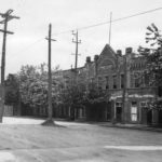 Black and white photograph of a large industrial brick building. There is a road in front of it with lots of wooden electrical poles and wires running alongside
