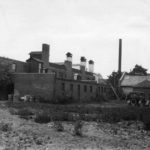 Black and white photograph of industrial brick buildings. Two vintage cars are seen at the right of the image