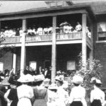 Black and white photograpj of a two storey brick building, with many people standing in front of the building an on the second storey porch