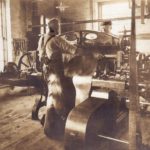Sepia photo of two men, standing at a machine, working with a large piece of leather