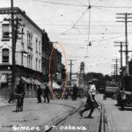 Black and white photo of a busy street, around the turn of hte 20th century. There are vintage cars and many people on the street. One building has been circled in orange.
