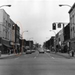Black and white photo of a streetcape, mid 20th century. There are cars and people on the sidewalks. A building has been circled in orange.