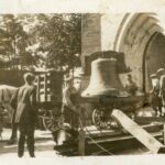 Sepia photo of a large bell being brought into a building by five men. There is a horse and carriage seen to the left of the image.