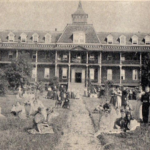 Two storey school building with several female students posed on the lawn out front of the building