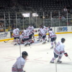 12 Oshawa Generals players skating near their net at an end of the rink. 2 are further out, going right. The rest are huddled near the goalie in net.