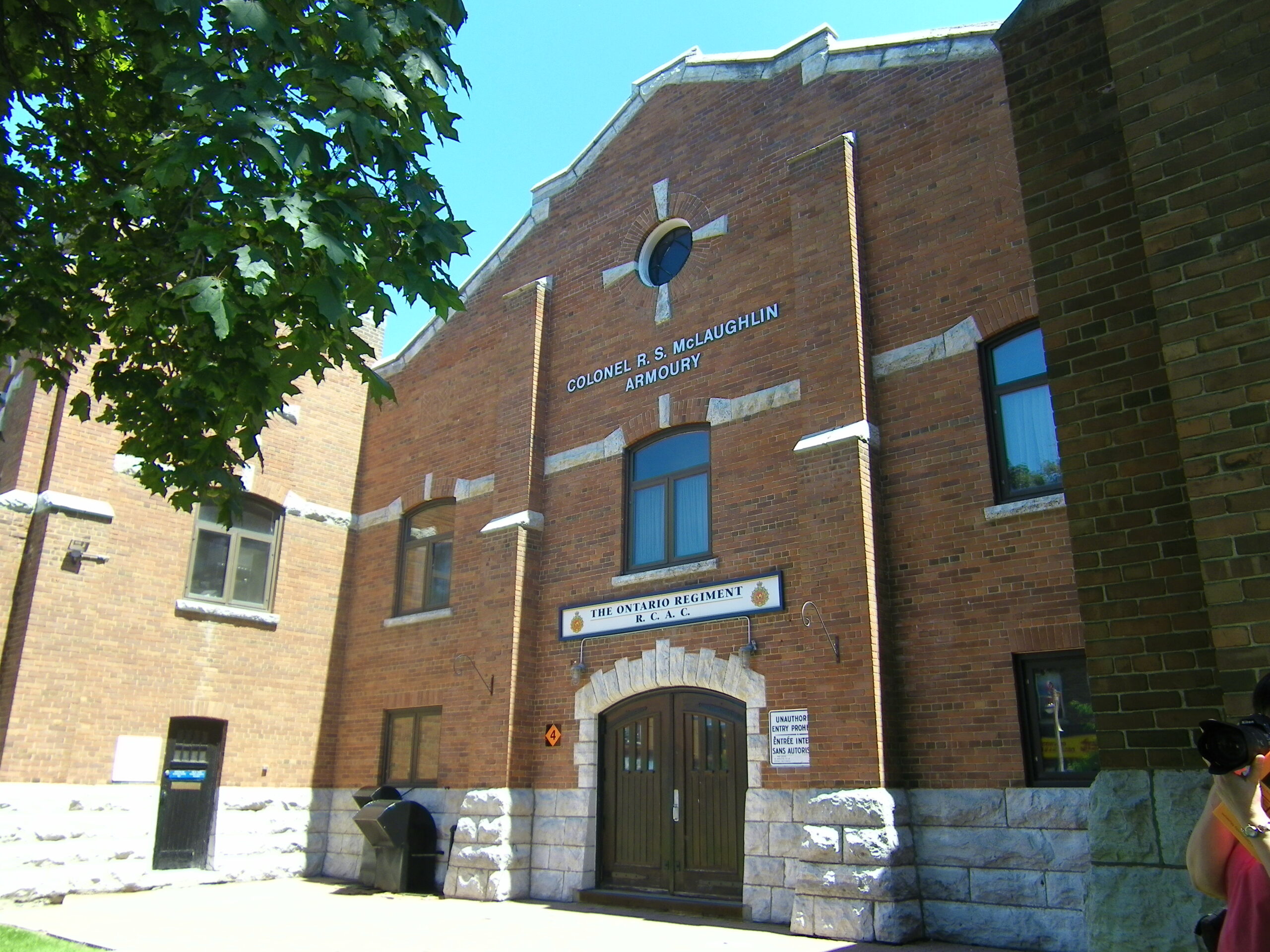 The entrance to the Col R.S. McLaughlin Armoury on Simcoe Street N, the image was taken from southwest of the building around 10 meters from the entrance. one of the two square towers attached to the building's entrance is covered from the camera view by a tree on the left side of the image.