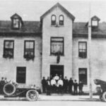A black and white image of the Commercial Hotel circa 1915. A group of 11 people are seen standing in front of the entrance to the building, which has two automobiles parked on the street, the one on the left side has two people in it.