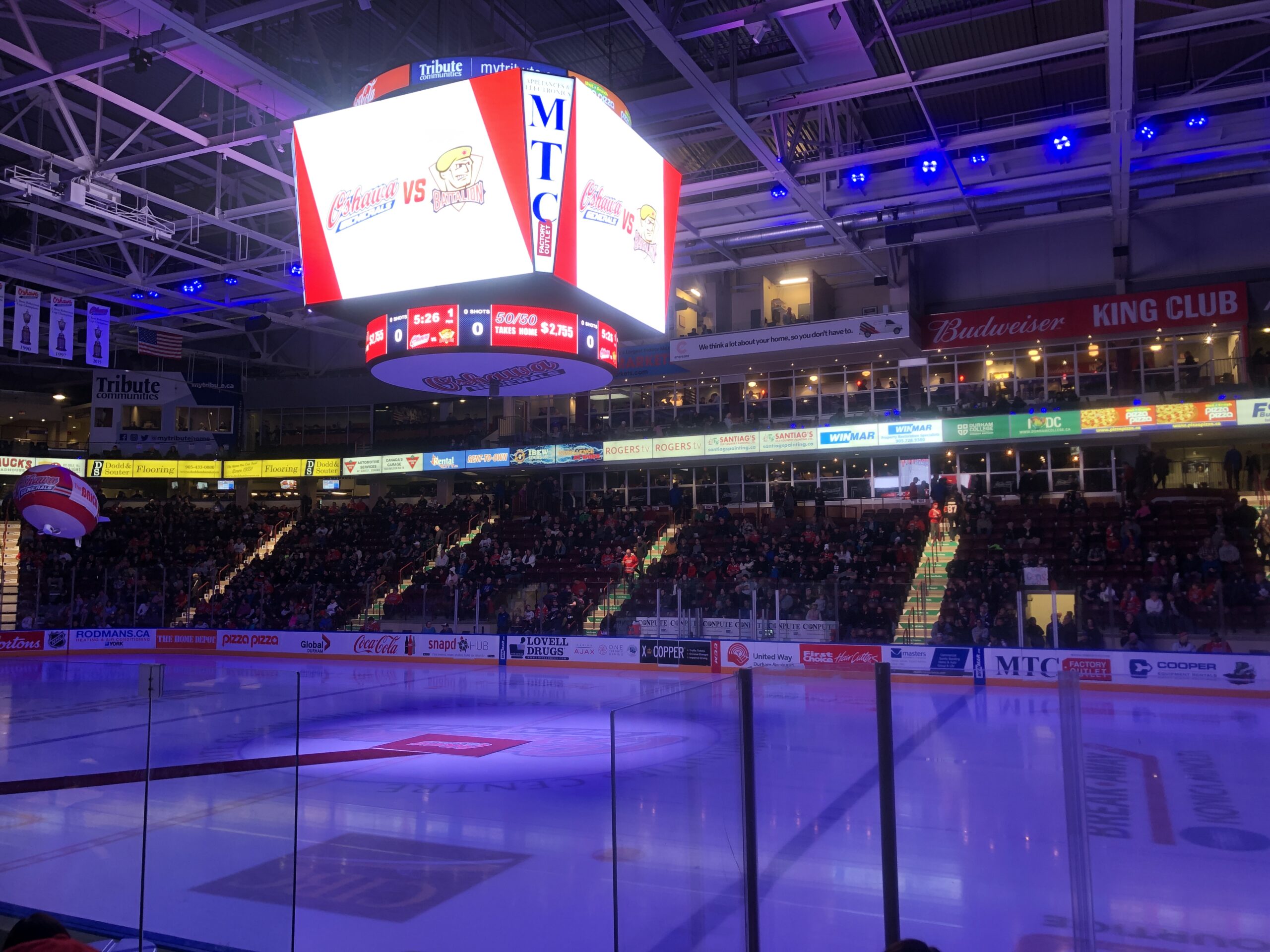 The interior of the TCC Arena as purple lights illuminate the rink. The crowd are getting to their seats, and the game will begin in five minutes after the photo was taken. The photo captures the opposite side of the rink from where it was taken.