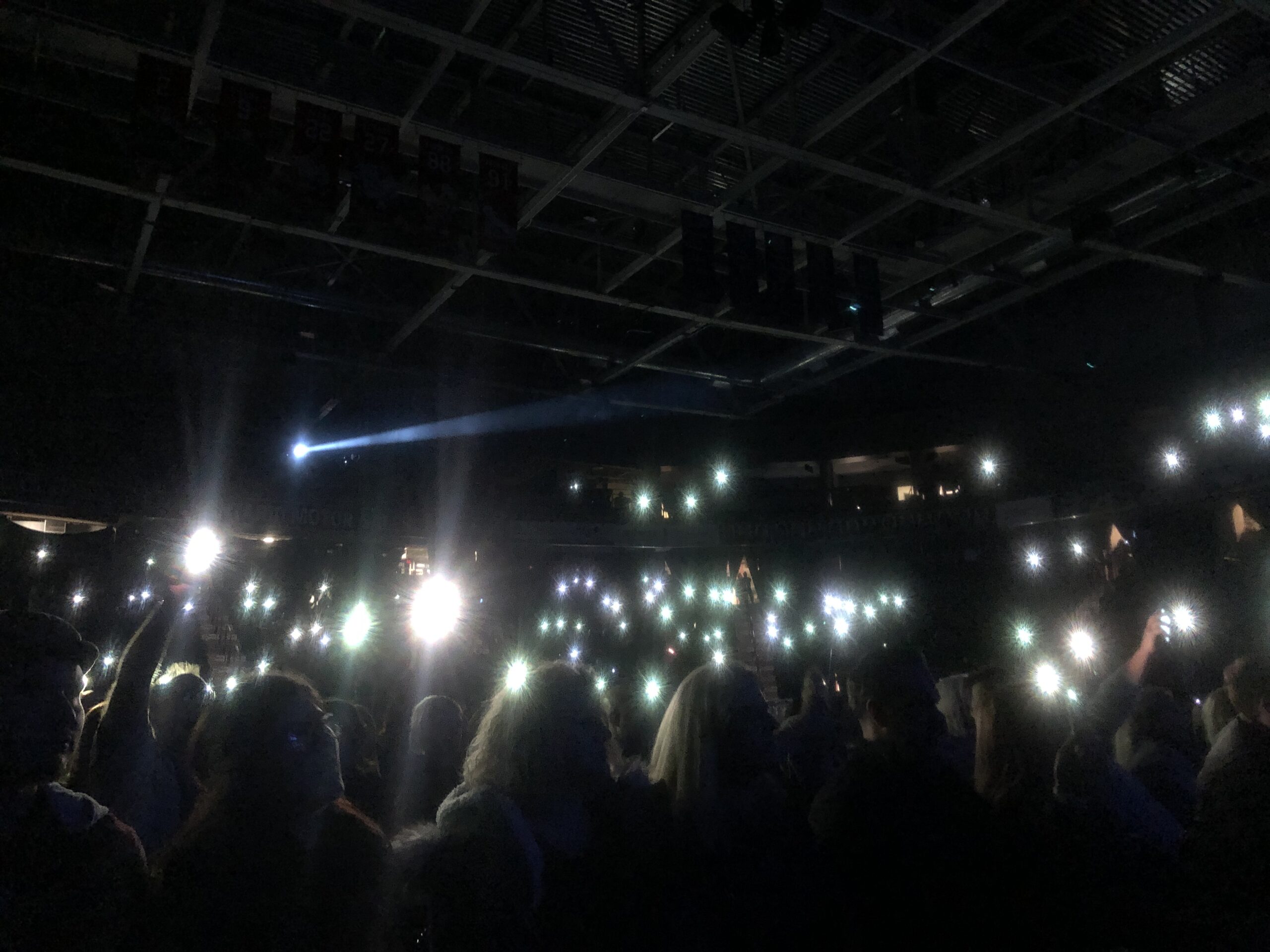 The darkened interior of the TCC during a performance by the musical rock experience We Will Rock You, a tribute musical to the band Queen. The audience have their phone flashlights on, swaying to the music.