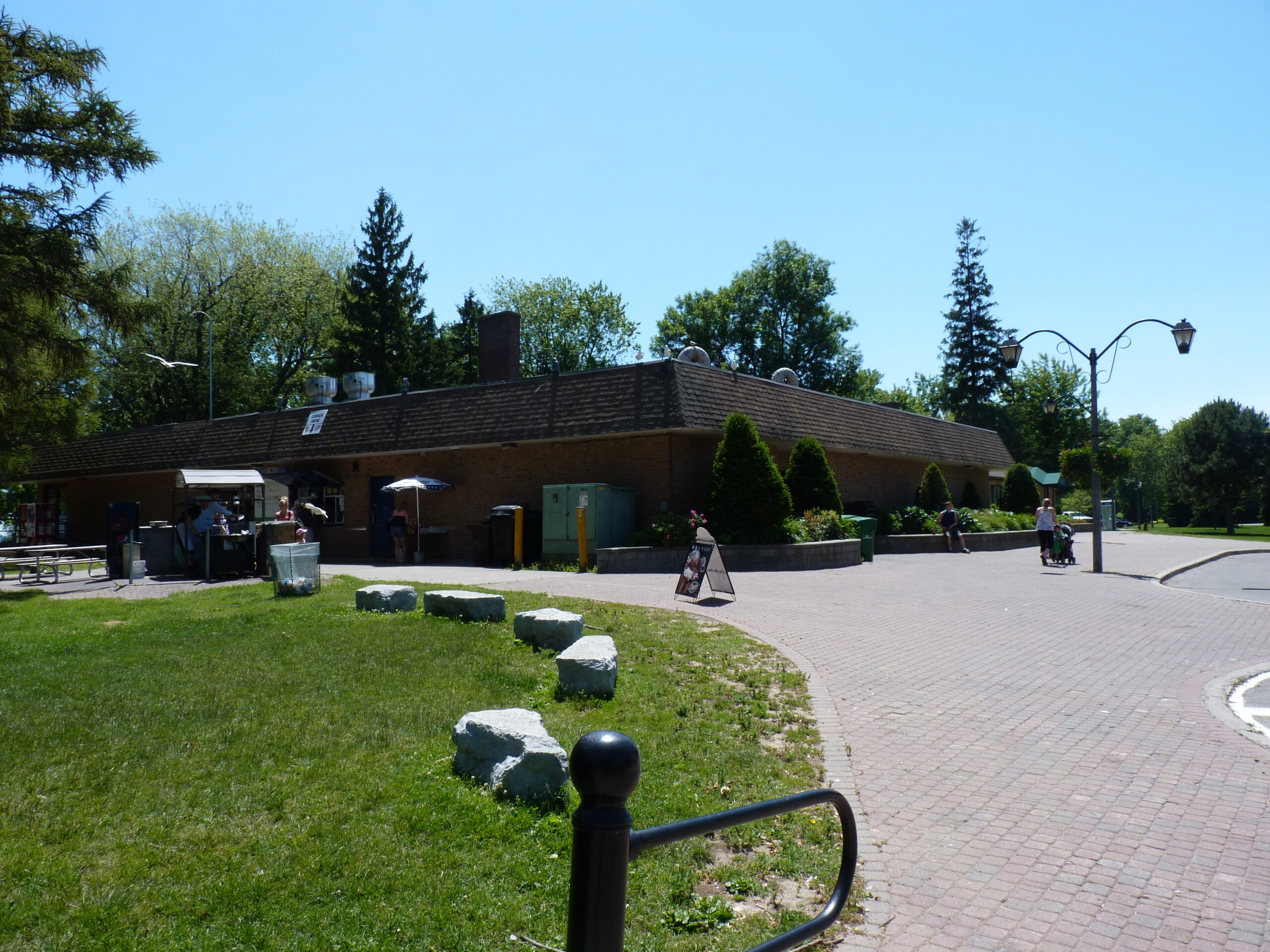 An image of a cloudless, sunny day at the Jubilee Pavilion circa 2010s, with the building's snack bar visible on the left side of the image. The image is facing west, from around 10 meters away from the building. The few people in the image are around the snack bar. There is a small sign advertising ice cream on the path heading towards the snack bar.