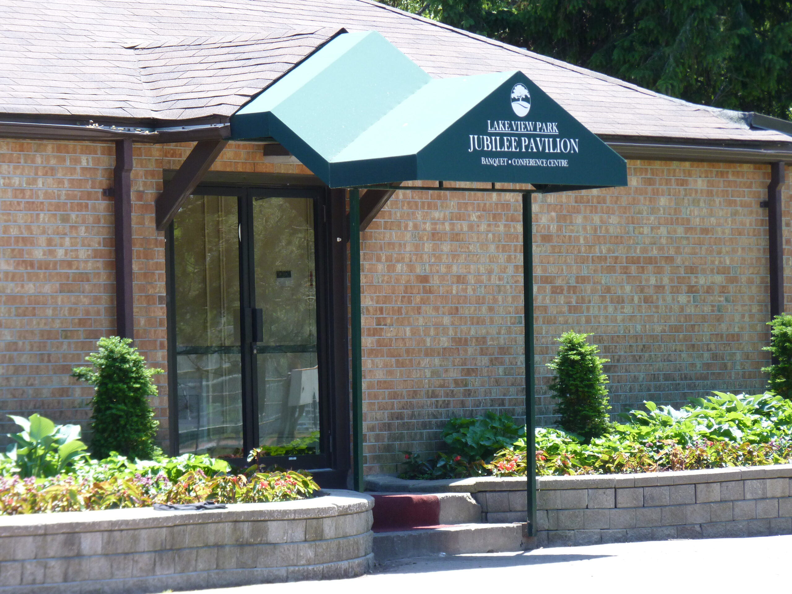 An image of the front entrance to the Jubilee Pavilion circa 2010s. There is a green metal hood over the entrance to keep the doorway shaded. There are also green plants growing in the small garden areas on either side of the door. The gardens are lined with little grey bricks serving as mini walls.