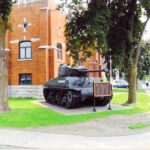 A tank is seen on display on the northwest side of the Col R.S. McLaughlin Armoury. It is a very sunny day, and the grass is very green as the image's brightness is high.