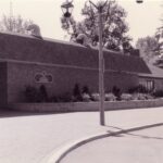 A black and white photo with a very slight warm tone of the Jubilee Pavilion building during the 1990s. Two lamp poles are visible in front of the main entrance and additional length of the building of to the left side.