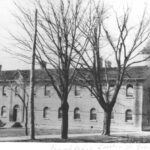 A black and white image of Centre Street School before 1924. The image shows the side of the school, with the bell tower on the right side of the image and the rest of the side length of the building taking up the rest of the image. There are a couple trees in front of the school, and none of them have leaves. Though the building and trees are the only visible thing in the image, the image was taken from approximately 40 meters away from the building.