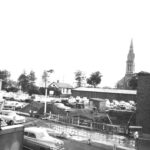 A black and white image of a view showing excavating work in progress for the Lancaster Hotel addition to what was the Commercial Hotel. There are multiple 50s era cars in the image, most parked around the construction site. A tall church can be seen in the background.