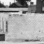 A black and white image of the Commercial hotel during construction efforts as it's name was being changed to the Lancaster Hotel. The image shows a small brick building with construction debris lined along the walls, and wooden planks left on the roof. There are two sections of wooden scaffolding on one side of the building where additional length is being added. The roofs of two houses can be seen in the background.