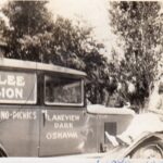 A slightly blurred black and white image of a man posing on the front hood of a 1930s era car with "Jubilee Pavilion" on the side. There are trees in the background and the image is slightly pointed upwards.