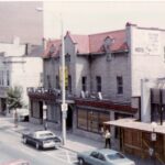 A view of the Lancaster Hotel during the 1970s taken from across the street on a slightly higher level than the second floor. The image shows the red roofed hotel, with three floors and an entrance in the middle of it's front wall. There are two cars parked on the street, and someone walking down the sidewalk, which is on the Lancaster's side of the road.