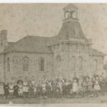 Large group of children posed in front of a brick school house. The building has rounded windows and a bell tower. The image is black and white.
