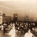 Picture of a classroom with students at their desks in rows and classroom settings in the background
