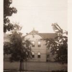Black and white picture of a two-story building seen across the street with trees covering parts of the sides and the inscription “Mary St. School”