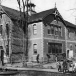 Black and white picture of a two storey building from the side with students outside the school entrance and bare trees blocking the view