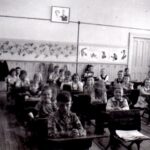 Black and white picture of children sitting at desks in rows smiling at the camera in a classroom