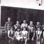 Black and white picture of two rows of children in front of a blackboard smiling at the camera