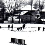 Black and white picture of a skating rink beside a building with students in pairs skating around on the ice and houses in the background