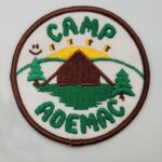 Colour photograph of a badge that reads Camp Ademac. It has a brown border, and in the centre is a brown cabin with green trees and a rising sun
