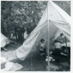 Black and white photo of a canvas tent with three people under it getting it set up