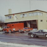 Rectangular building made out of brick with four garages, all with glass panel doors. One red truck and car are parked in the driveway.