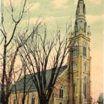 Colourized photo of a yellow brick building with ornate windows and a tall spire