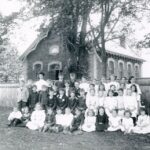 Black and white photo of a group of people posed in rows for the picture. They are in front of a wood fence. On the other side of the fence is a brick building, with a sign reading "School Section No. 4, East Whitby, Erected 1874"