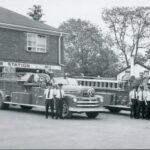 Black and white photograph of a rectangular brick building that is partly painted in white. Two trucks are displayed with firemen standing beside them.