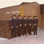 Five firemen in black suits standing in front of a brick building with the words “fire station 3” put up on the wall.