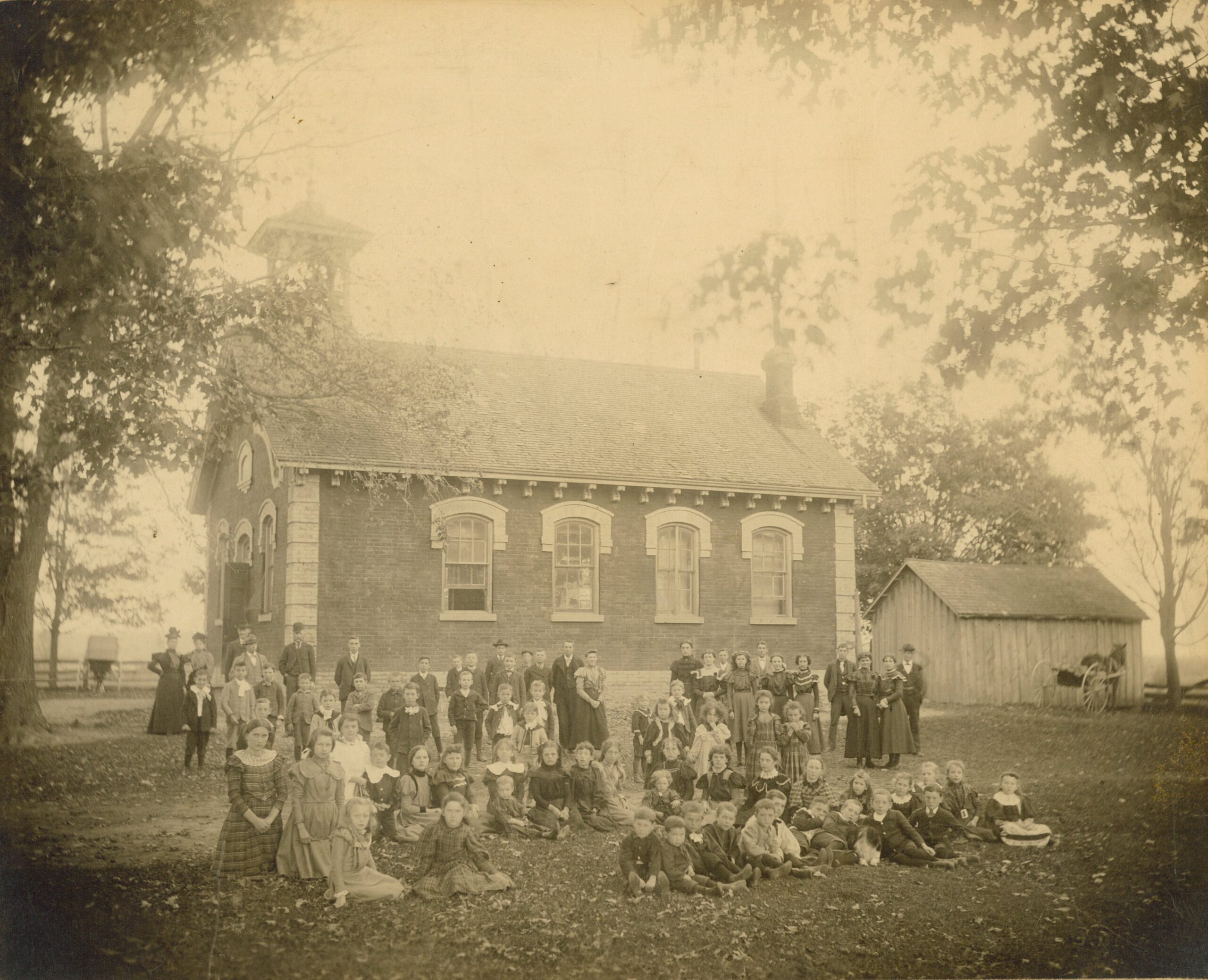 Sepia photograph of a group of people posed for the picture. They are in front of a brick building, and there are trees to the left and right of the image.