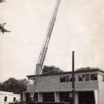 Black and white photograph of a rectangular brick building. There is a dispatch ladder truck in front of it.