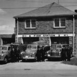 Black and white photograph of a rectangular brick building that is partly painted in white. Three trucks are displayed with firemen standing beside them.