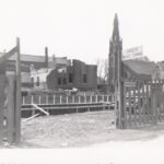 Black and white photo of a construction site. There are buildings, including a church, in the background