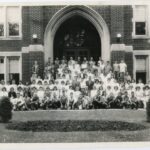 Black and white photo of a group of students and teachers out front of a brick building with a large archway