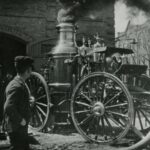 Black and white photograph of a vehicle that has large wheels and smoke coming out of the top.