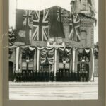 Black and white photo of a number of men, lined up outside of a two-storey brick building decorated with flags and a welcome sign.