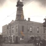 Black and white photo of a large, two storey brick building. The building has a clock tower, which is on fire. There is a fire fighter on the roof, and there is a crowd of people watching the scene