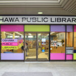Landscape photo of a glass wall paneled building with banners of purple, pink, and yellow decorating it. A large white banner, saying “Oshawa Public Libraries,” is placed at the top.