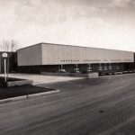 Black and white landscape photograph of a long rectangular building. The pavement and paved grass is clean. Large windows are stationed on all sides of the building.