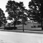 Black and white angled landscape shot of a long rectangular building, positioned behind three visible trees. Windows are placed on the sides of the building along with bushes on certain sides. The pavement and paved grass is neat.