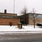 Photograph of a rectangular building positioned behind two visibly bare trees. Windows are placed on the front side of the building. Snow is neatly piled on the paved grass. Letter placed on the building read, “Central Collegiate Institute.”