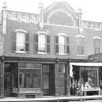 Black and white photo of a two storey building. There is a man standing in front of a doorway to the building